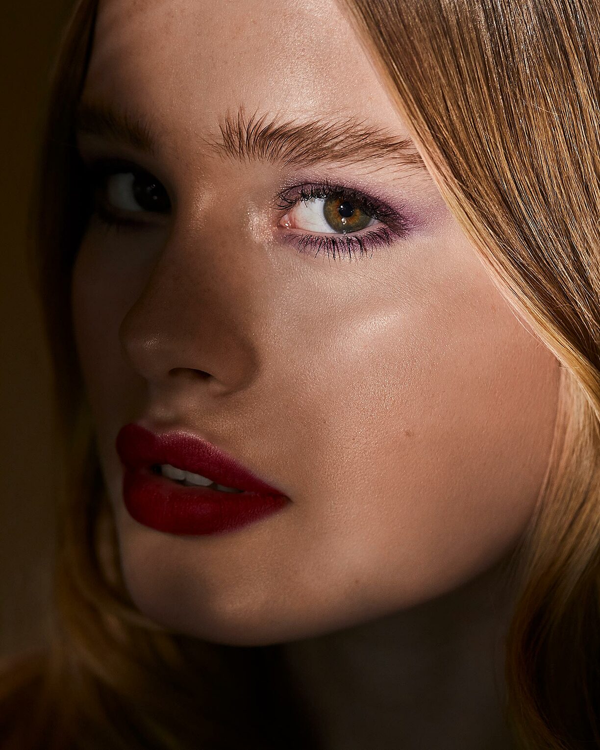 A beauty close-up with a blonde model. She wears red lips and a light purple eye shadow. A shadow is around her nose