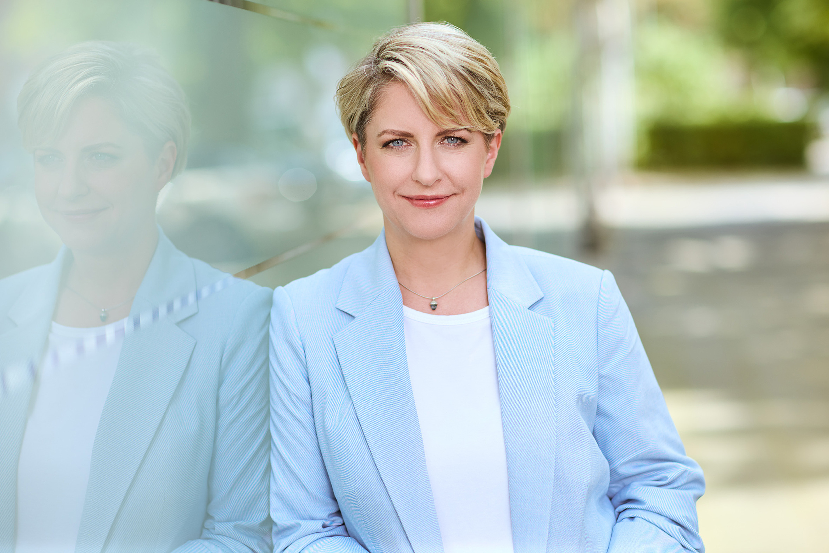 Unternehmerin steht draußen vor einer sich spiegelnden Wand in einem weissen Tshirt mit einem hellblauen Blazer. Grüne Bäume stehen um sie in der Unschärfe herum